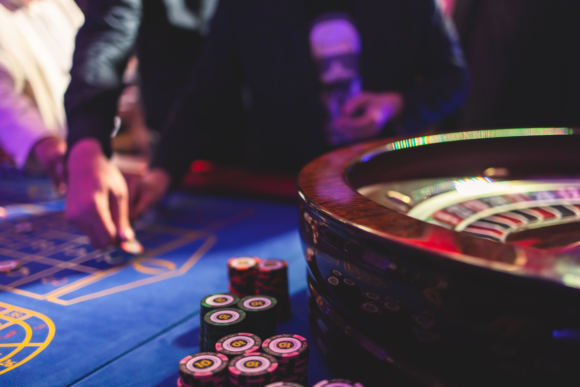 Vibrant casino table with roulette in motion, with casino chips, tokens, the hand of croupier, money and a group of gambling rich wealthy people playing bet in the background